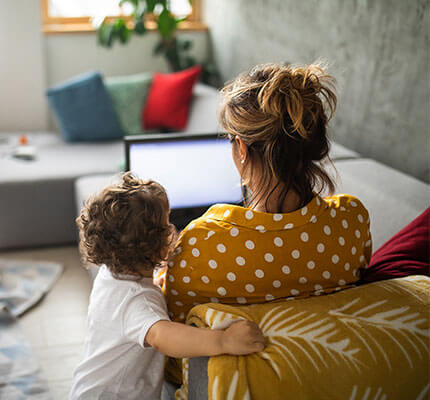 Over the shoulder view of woman on couch with a laptop as toddler looks on