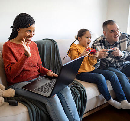 Woman sitting on a couch with her laptop while father and daughter play video games